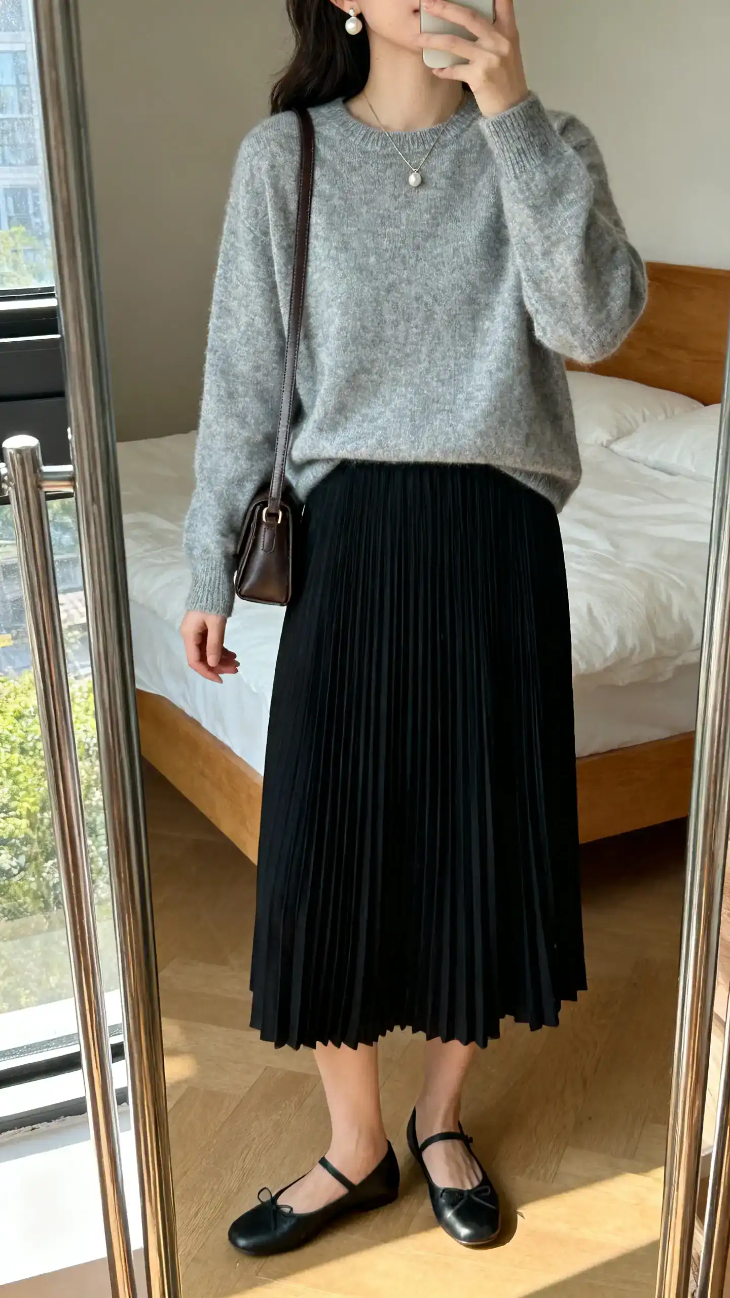 iPhone mirror selfie of a woman wearing a soft gray cashmere crewneck with a black pleated midi skirt, ballet flats, small leather shoulder bag, pearl earrings and a dainty pendant, face not visible, cropped at shoulders, bedroom mirror, morning window light, iPhone photo quality.