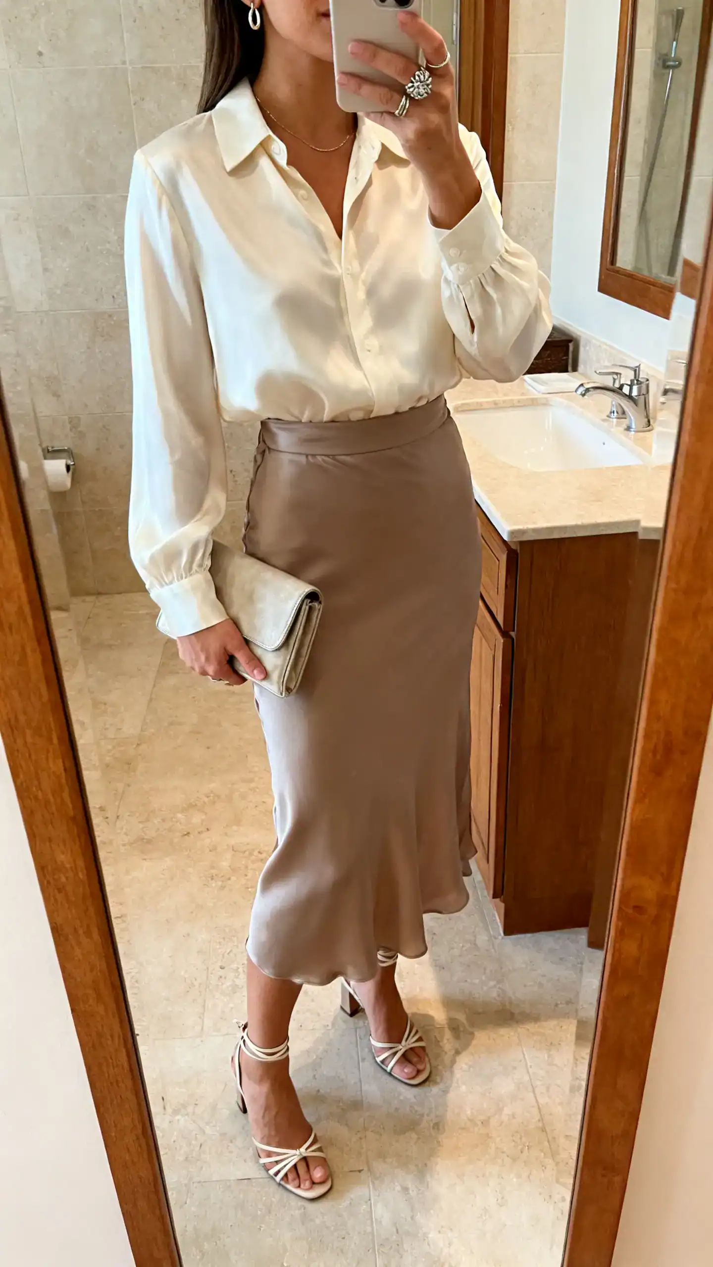 iPhone mirror selfie of a woman wearing an ivory silk blouse tucked into a matching taupe midi slip skirt, strappy low heels, compact clutch, statement ring and fine drop earrings, face not visible, cropped at shoulders, bathroom mirror, warm indoor light, iPhone photo quality.