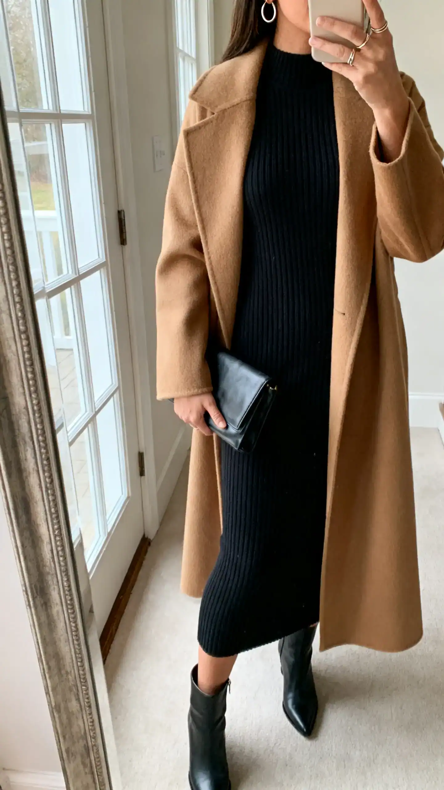 iPhone side-angle mirror selfie of a woman in a black knit column dress with a camel long wool coat, sleek ankle boots, structured clutch, fine hoop earrings and delicate ring stack, face not visible, cropped at neck, entryway mirror, natural window light, iPhone photo quality.