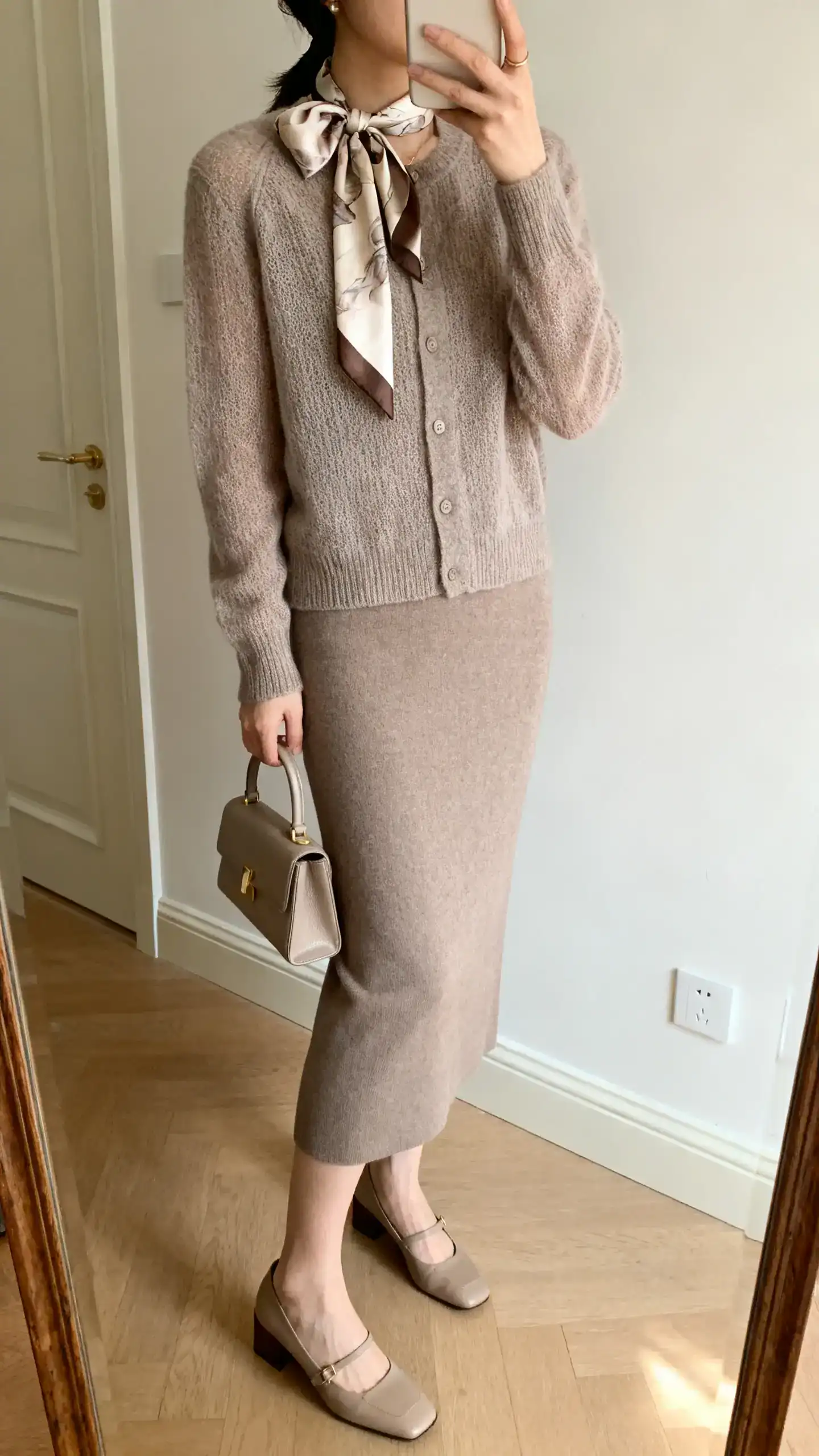 iPhone side-angle mirror selfie of a woman wearing a silk scarf tied at the neck, a fine knit cardigan in soft taupe, and a matching column skirt, with low block-heel pumps and a small top-handle bag, face not visible, cropped at shoulders, soft indoor lighting, iPhone photo quality.