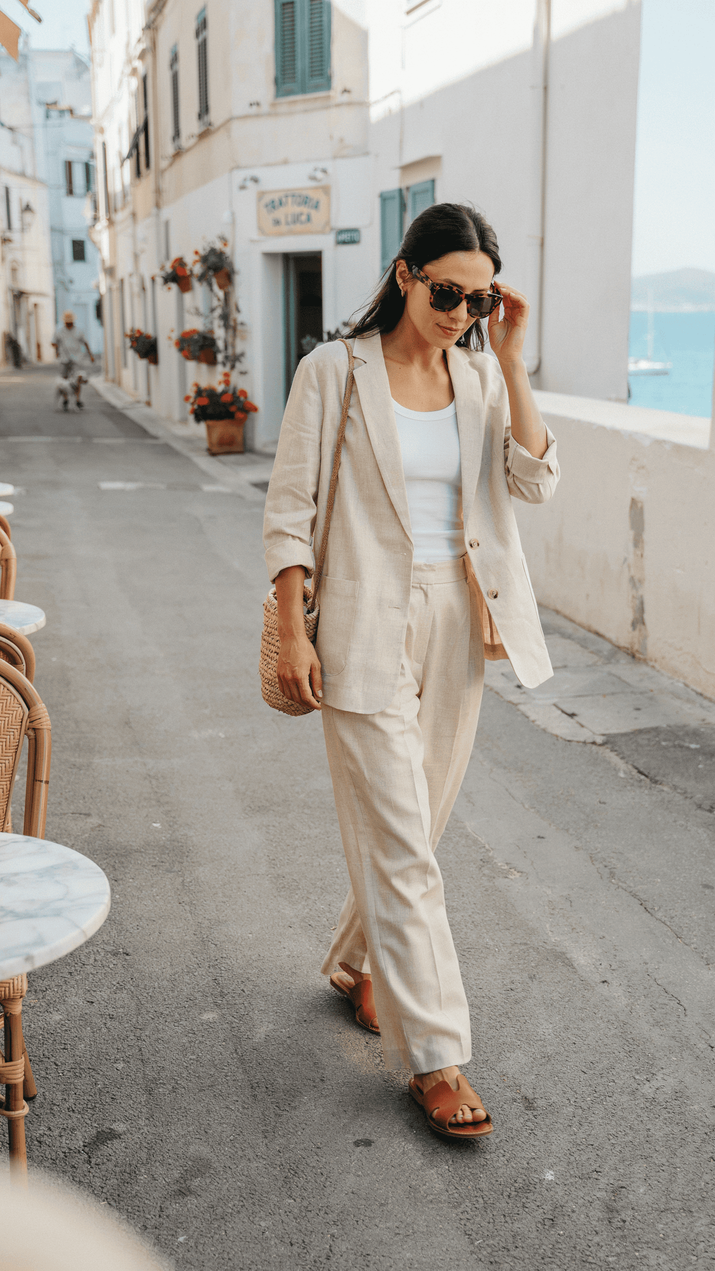 Woman wearing a relaxed beige linen suit over a white tank top with leather sandals and oversized sunglasses.