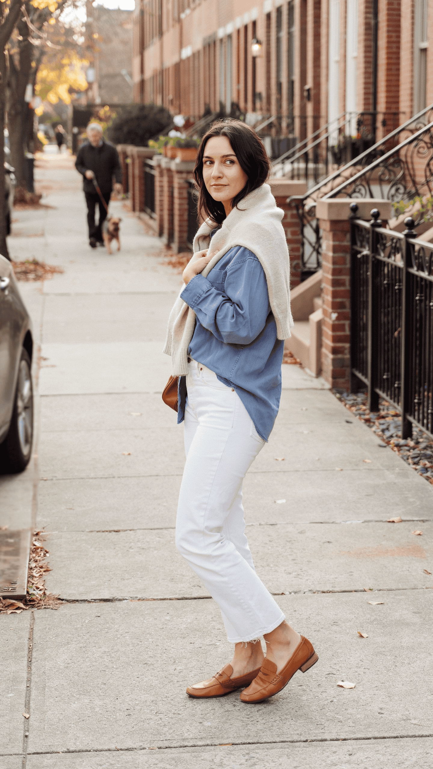 Woman wearing white jeans with a blue shirt and an ivory cashmere sweater draped over her shoulders.