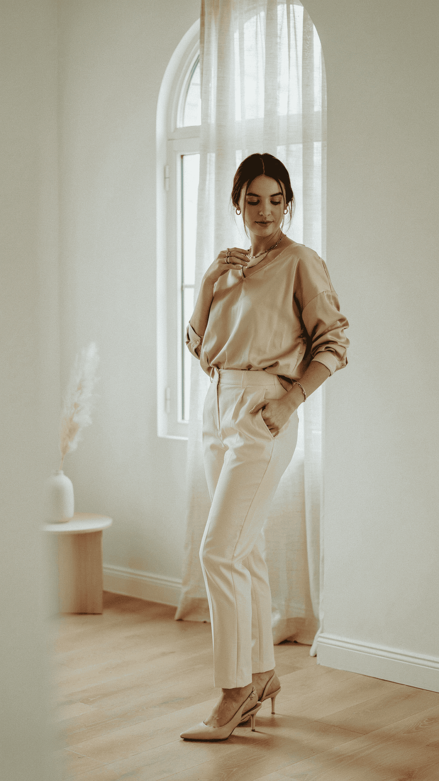 Champagne silk blouse tucked into cream high-waisted trousers with nude heels and gold jewellery.