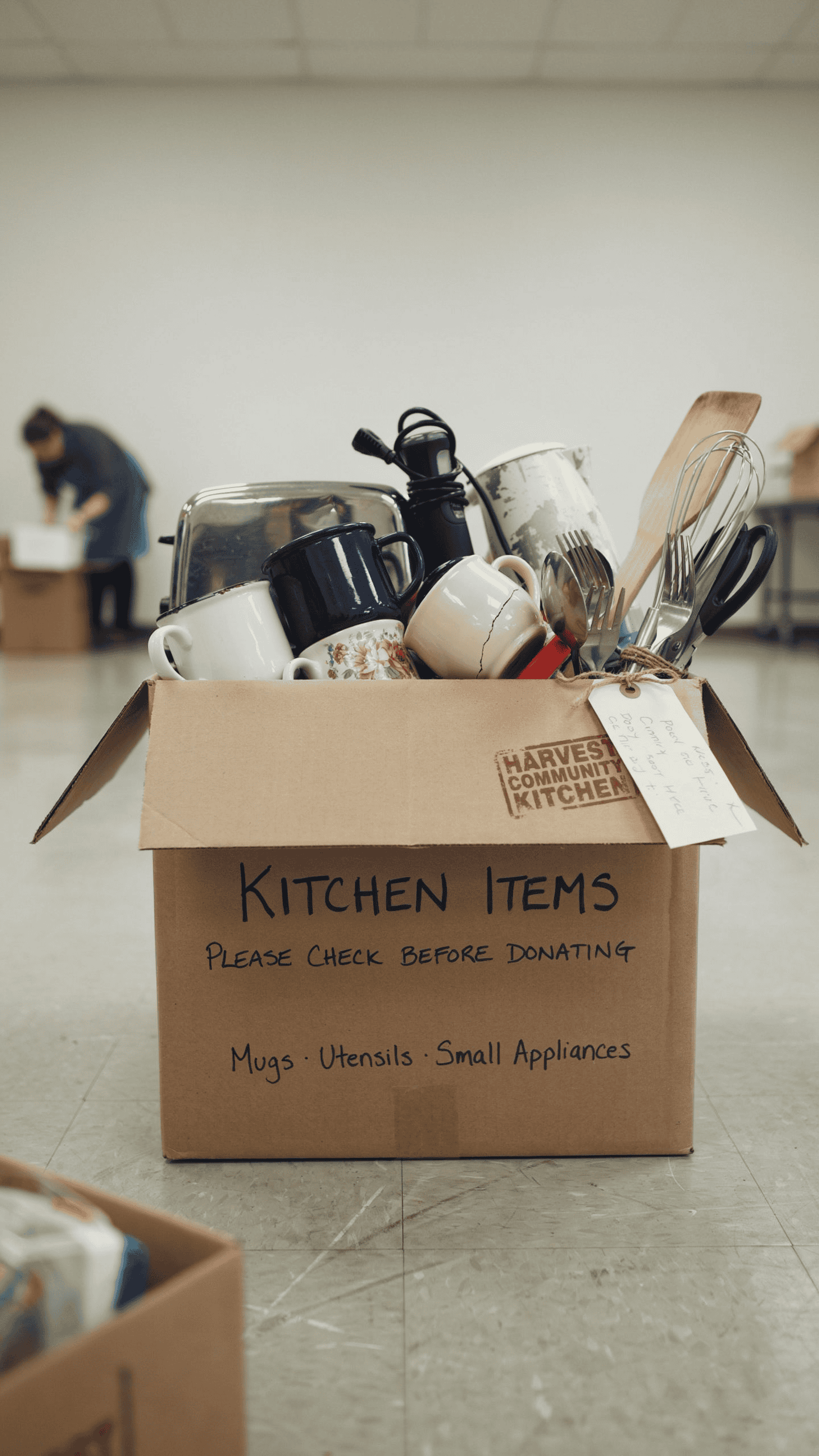 Cardboard donation box filled with extra mugs, utensils, and small kitchen appliances.