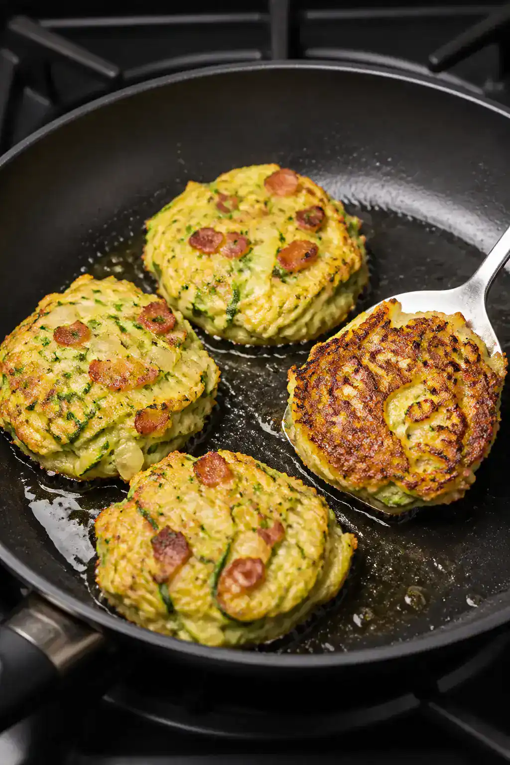 Cooking process: Zucchini and bacon breakfast fritters frying in a skillet, four patties gently flat