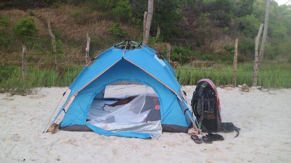 Blue camping tent set up on a sandy beach in El Nido with green grass and trees in the background. A backpack and sandals are placed next to the tent, suggesting a camping trip or beach outing before embarking on boat tours in the stunning natural setting of Palawan.