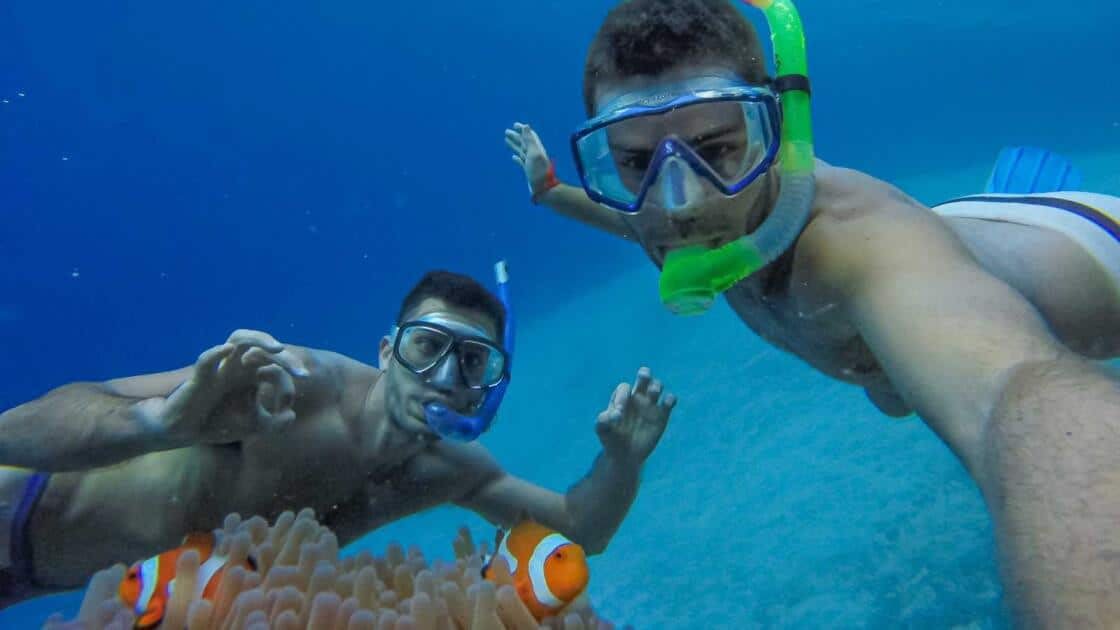 Two people snorkeling underwater with masks and snorkels take a selfie near clownfish swimming around a sea anemone. They're gesturing towards the fish, surrounded by Palawan's crystal-clear waters—a perfect snapshot of their island hopping adventure.