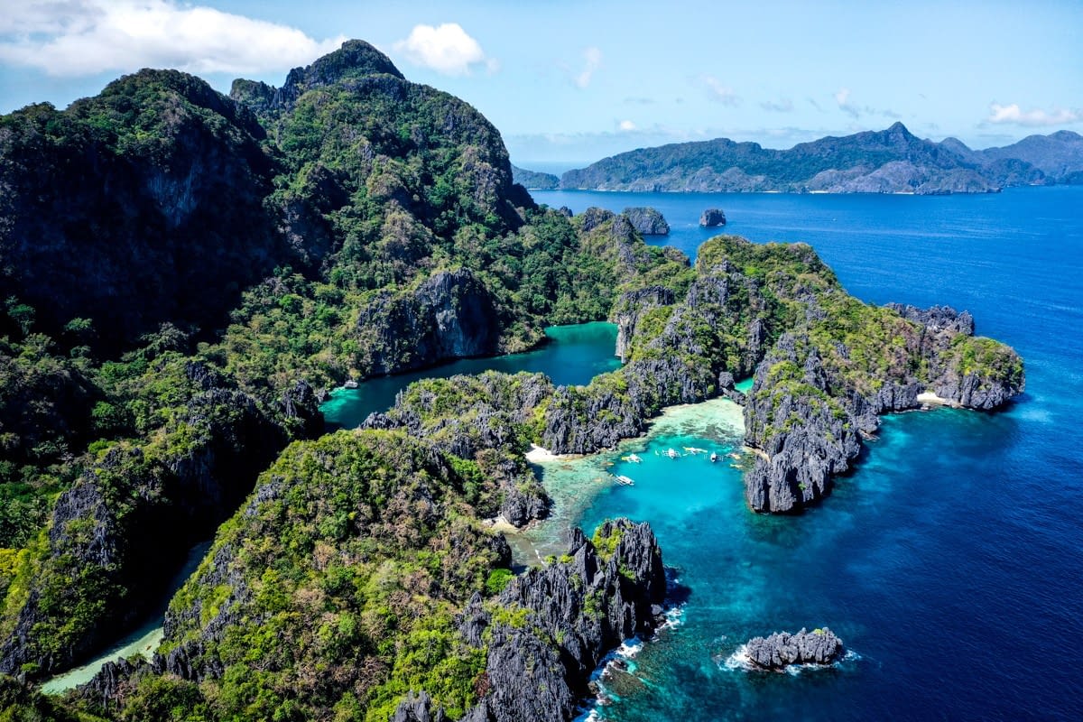 A stunning aerial view of Palawan reveals a lush, mountainous island surrounded by vibrant blue ocean waters. The landscape features rugged cliffs, green foliage, and small inlets with turquoise lagoons. Distant islands are visible under a clear blue sky, perfect for El Nido's famous boat tours.