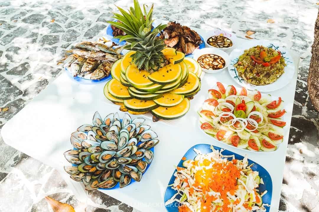A white table outdoors is filled with a variety of dishes, including sliced watermelon with a pineapple on top, grilled fish, mussels, guacamole, sliced tomatoes with onions, and a salad. The table is set on a stone patio in Palawan, offering an idyllic taste of the Philippines.