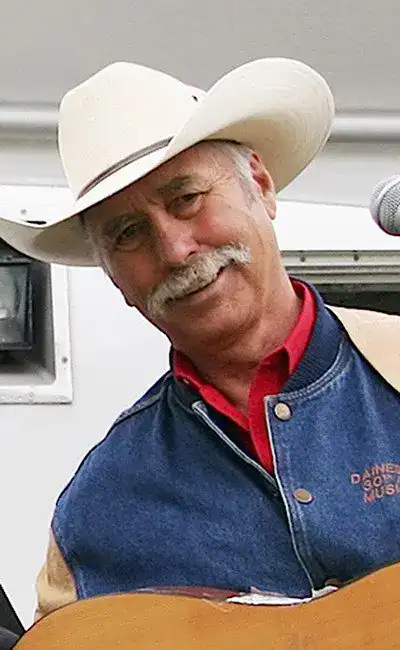 A cowboy musician smiling with a hat and guitar at MooseMedia, showcasing live country music performances.