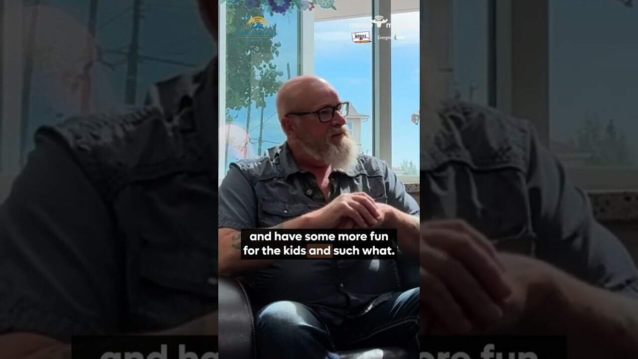 Relaxed man wearing glasses and a vest sitting in a bright indoor space with large windows.