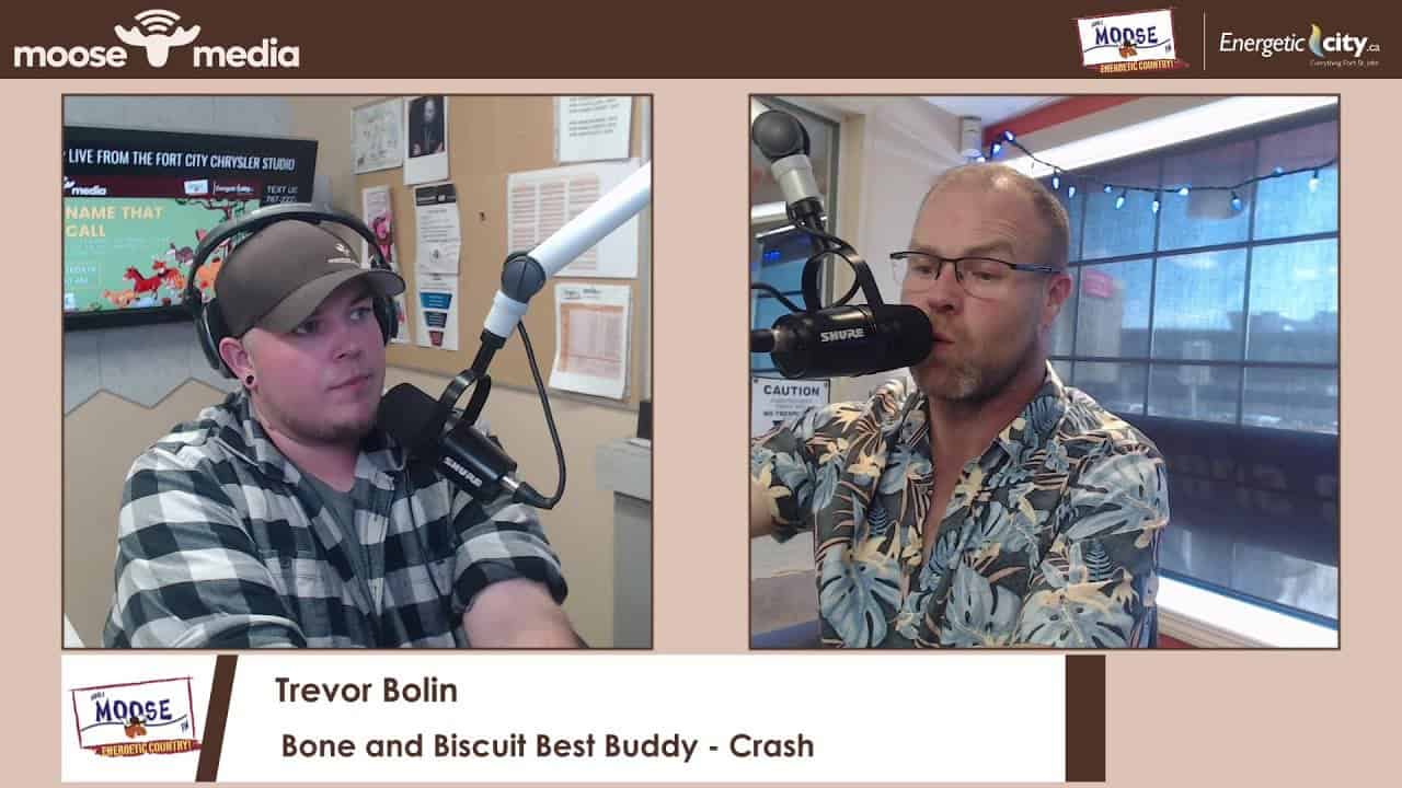 Young man in plaid shirt with headphones speaking into a microphone in radio studio.