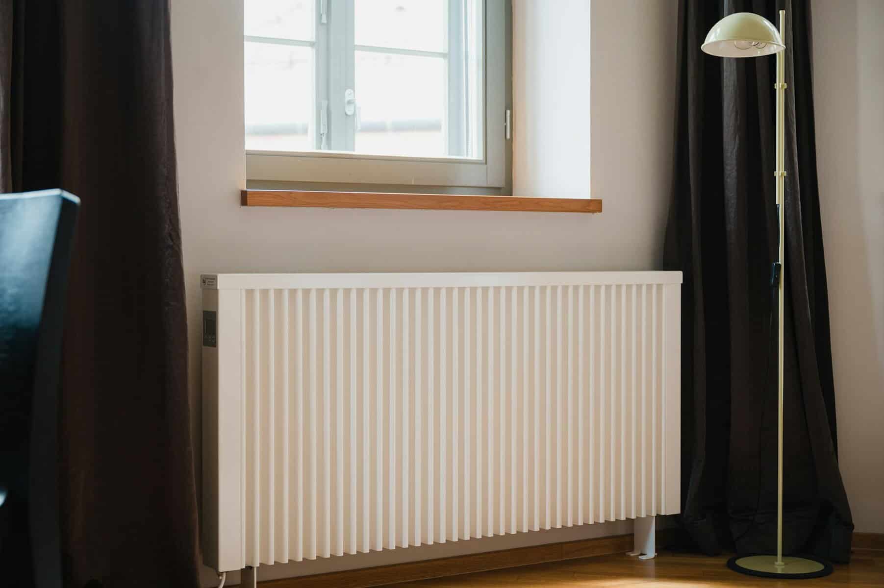 Efficient home heating radiator under window with black curtains and floor lamp, showcasing professional HVAC services for optimal indoor climate control.