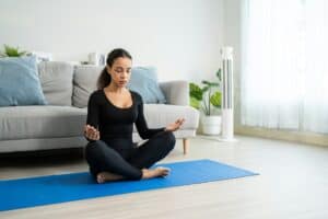 RELAXING WOMAN MEDITATING IN LIVING ROOM FOR STRESS RELIEF, HEALTHY LIFESTYLE, AND MINDFULNESS, WITH AN AIR PURIFIER IN BACKGROUND.