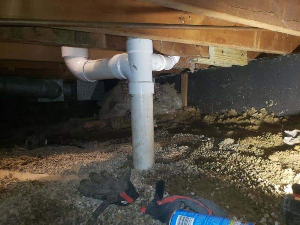 A white PVC cleanout and drain fitting installed on the sewer line in a crawlspace beneath floor joists, gravel and soil below