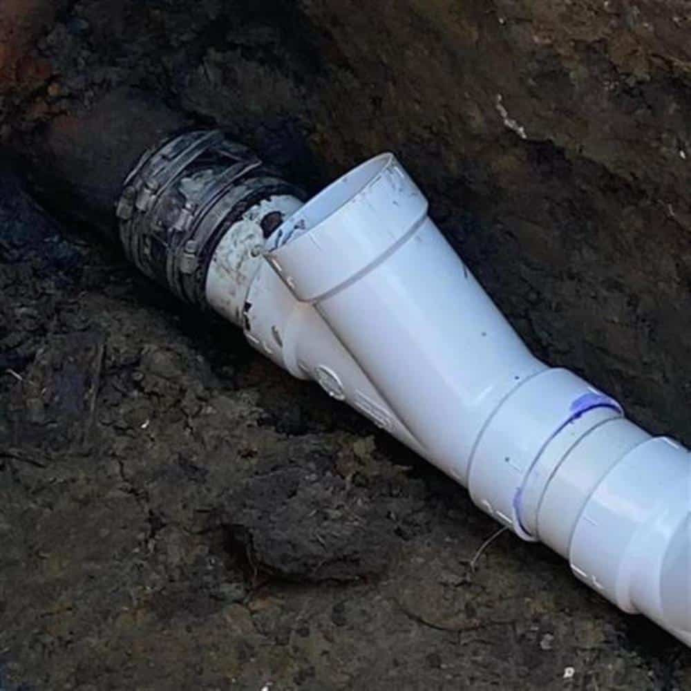 Close-up of a new white PVC sewer fitting joined to the old pipe with a stainless-banded flexible coupling, sitting in wet dark soil at the bottom of a trench