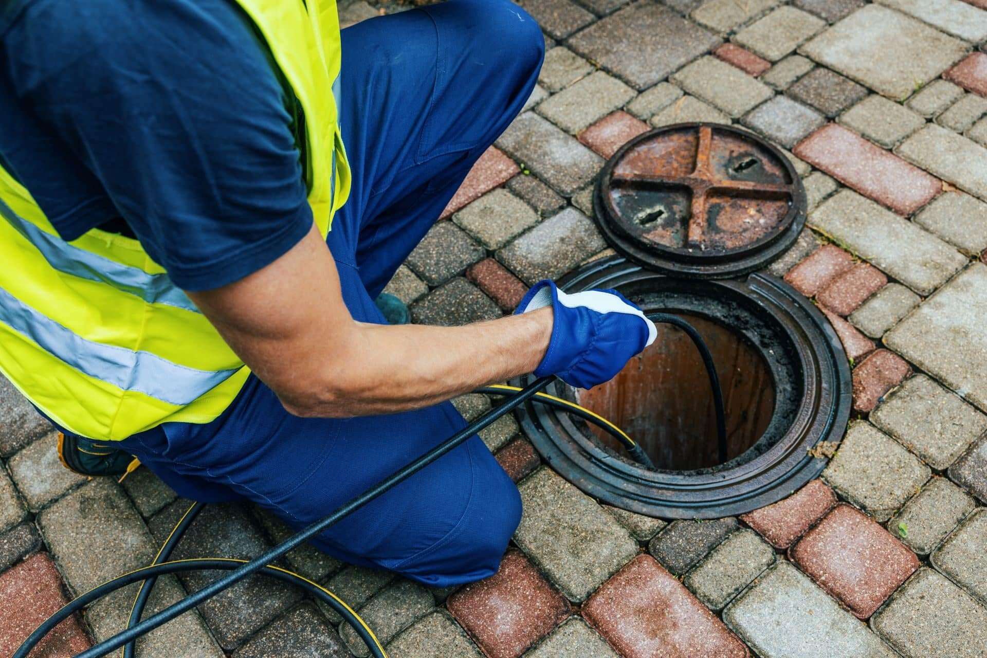A worker in a reflective vest kneels on paved bricks, inserting a cable into an open manhole for Trenchless Sewer Repair or inspection—an innovative, no-dig solution often handled by a plumber near me.
