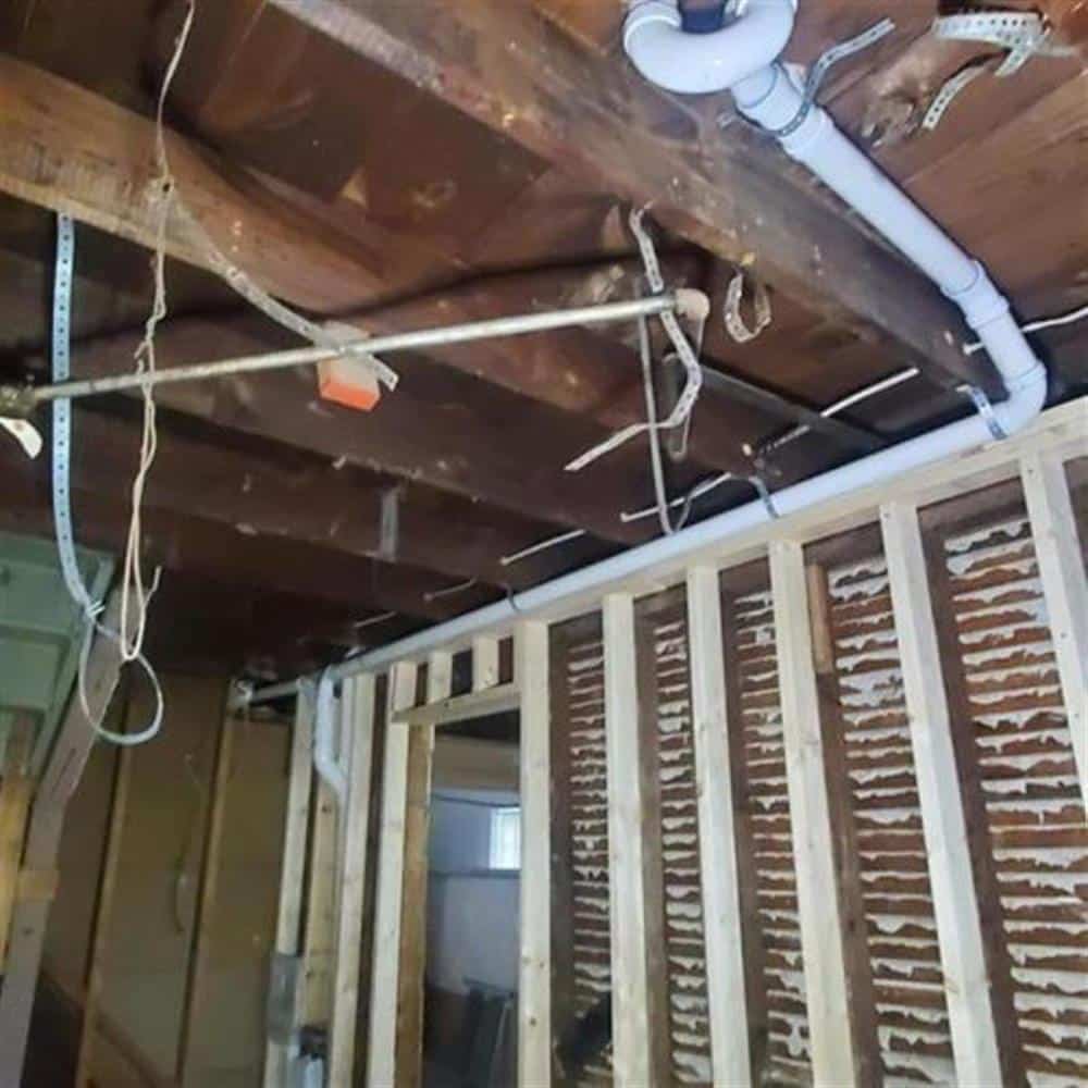A new white PVC drain line running the length of a basement ceiling between old floor joists above exposed lath-and-plaster walls