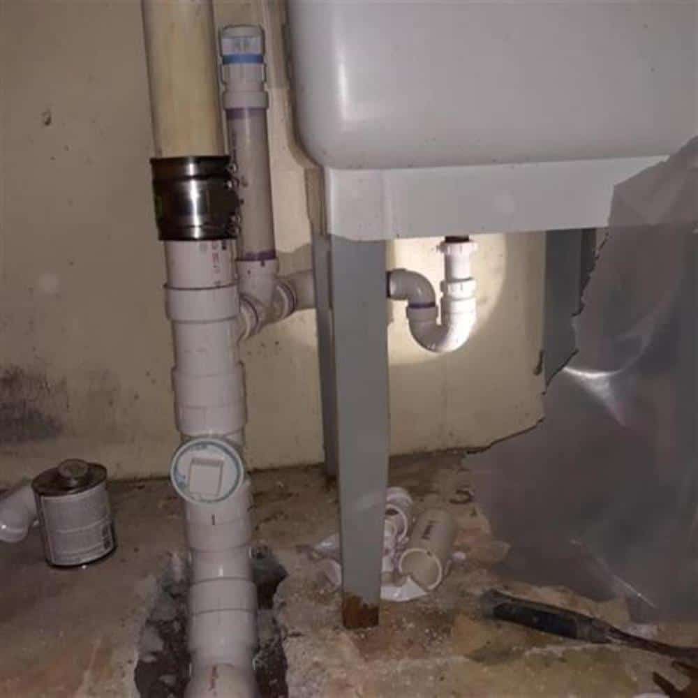An older basement where a cast-iron drain stack has been transitioned to new white PVC with a shielded coupling beneath a laundry tub