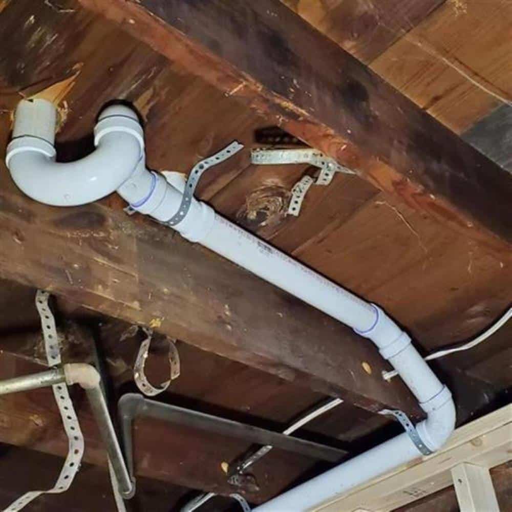 A new white PVC drain line with a long-sweep elbow, hung from floor joists with perforated metal strapping in an older basement