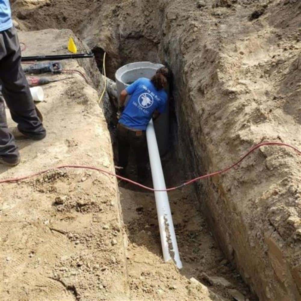 A Bison Plumbing technician working at the bottom of a deep open trench, setting a new white PVC sewer lateral with a red safety line running along the pipe