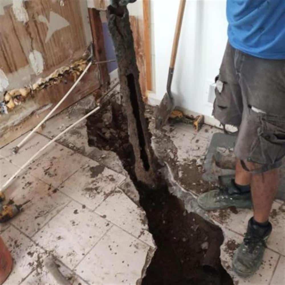 An old sewer drain line being dug out of a narrow trench cut through a tiled floor in an older home, corroded pipe and dark soil exposed, a Bison Plumbing technician's boots and a shovel beside it