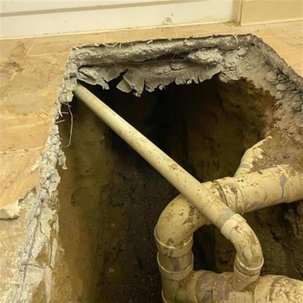 An aged sewer lateral with a joint and fitting exposed at the bottom of a deep hand-dug pit beneath a concrete floor on a Bison Plumbing repair