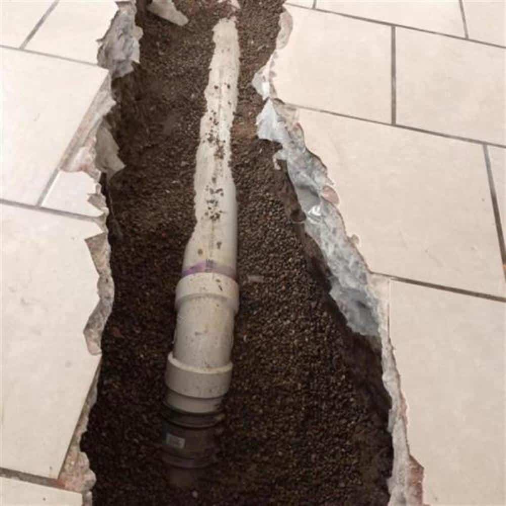 A trench cut across a tiled basement floor with a PVC drain line bedded in gravel, running toward the sump pit on a Bison Plumbing installation