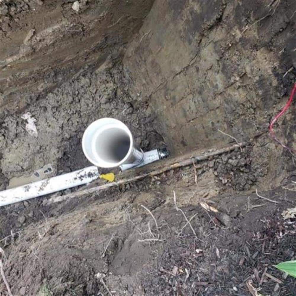 Looking down into a compact access pit at a new white PVC sewer fitting joined to the lateral with a banded coupling, dark backfill soil piled around the rim