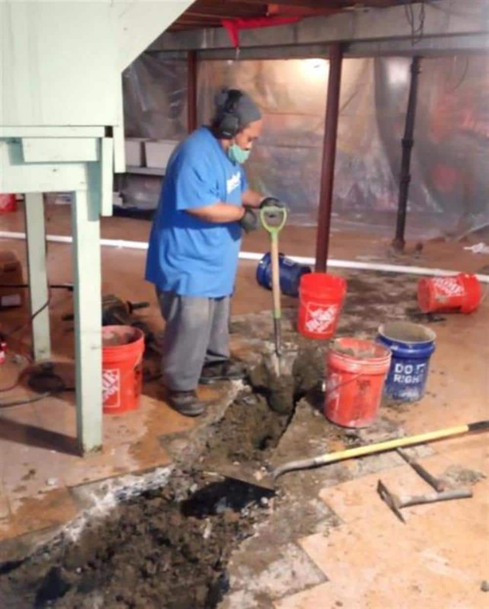 A Bison Plumbing technician opening a basement floor with a shovel to set a sump pit and drain line, buckets and plastic sheeting around the work area