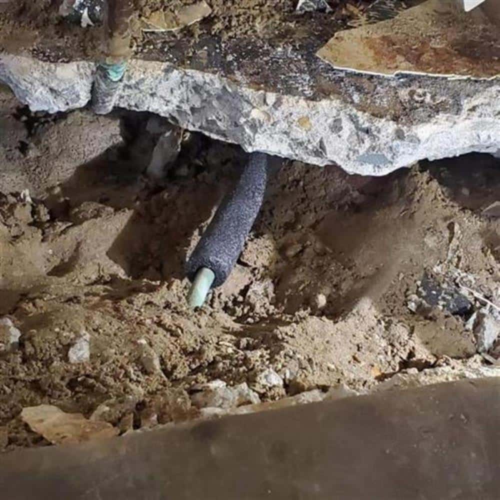 A sewer pipe exposed in the soil beneath a broken-out section of concrete basement slab during an under-floor sewer repair