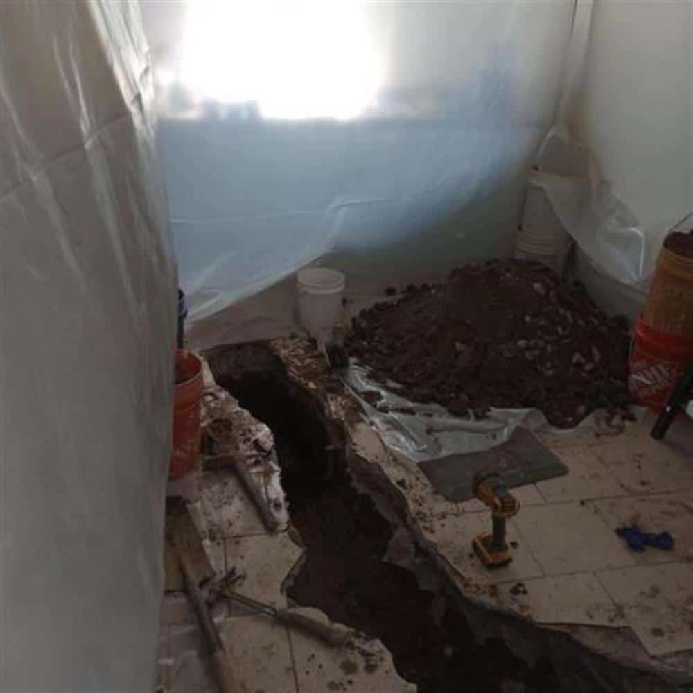 A basement in an older Oakland County home with the concrete floor cut open along the sewer line, excavated soil piled on plastic sheeting beside the trench