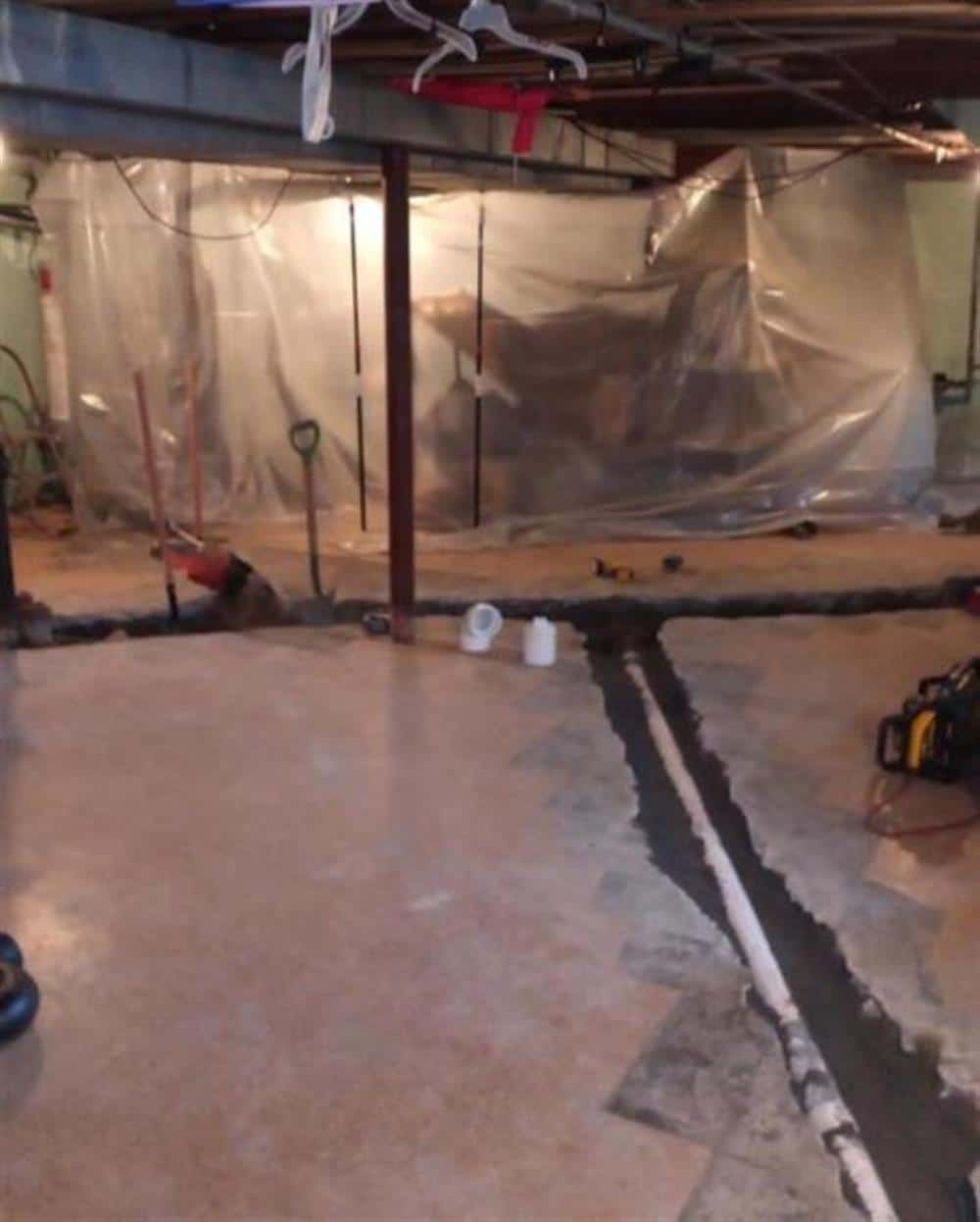 A finished basement floor cut open along the wall for a new drain line and sump system, with plastic sheeting protecting the room, on a Bison Plumbing job