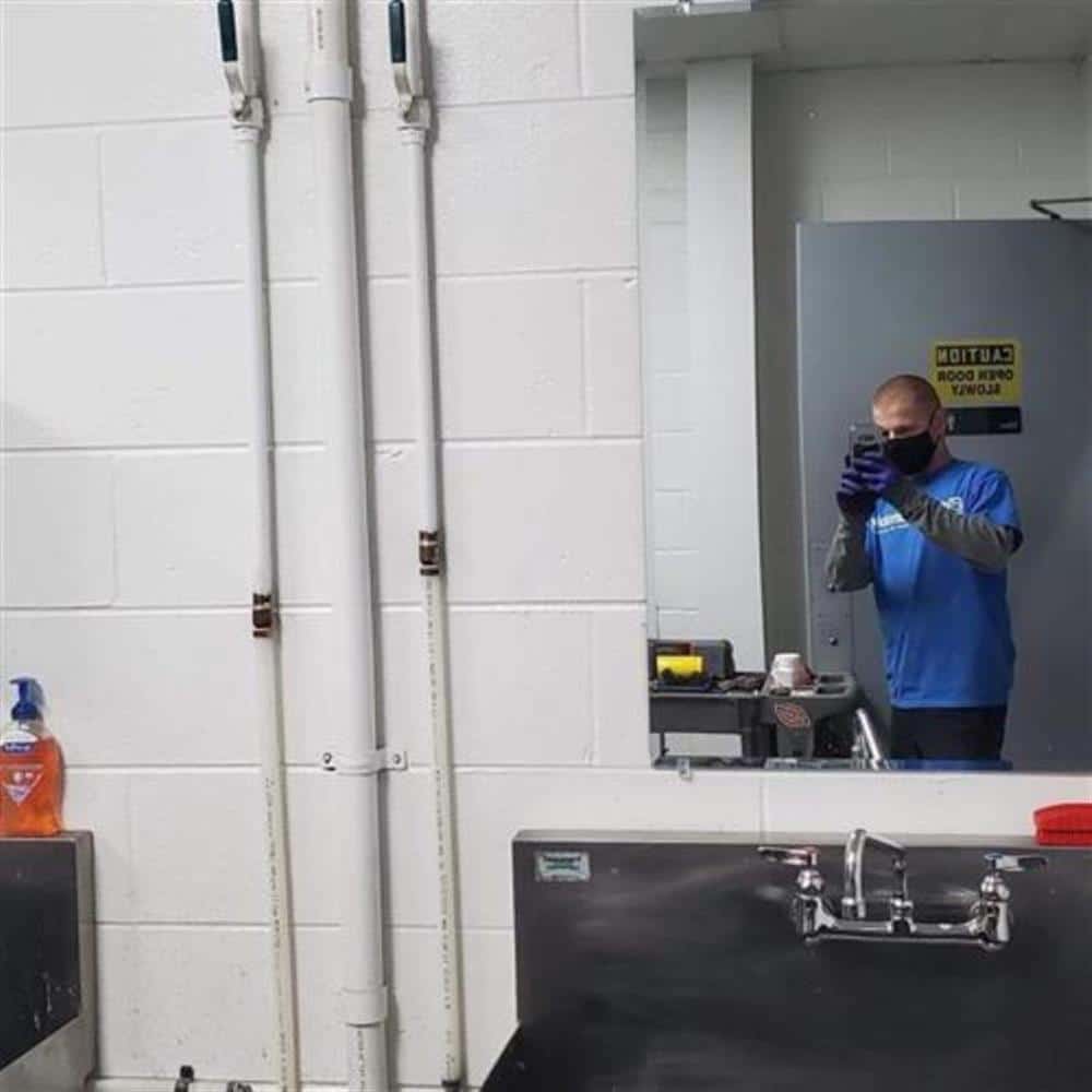A person wearing a mask and gloves is taking a photo in a mirror in an industrial-looking room with white walls, a sink, cleaning supplies, and a Caution sign on the door—possibly where plumbers in Warren MI work.