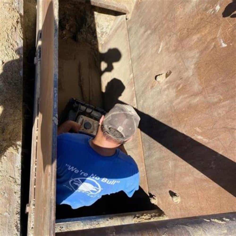 A Bison Plumbing technician in a 'We're No Bull' shirt working at the bottom of a shored access pit, plywood shoring on either side and sunlight casting his shadow on the wall