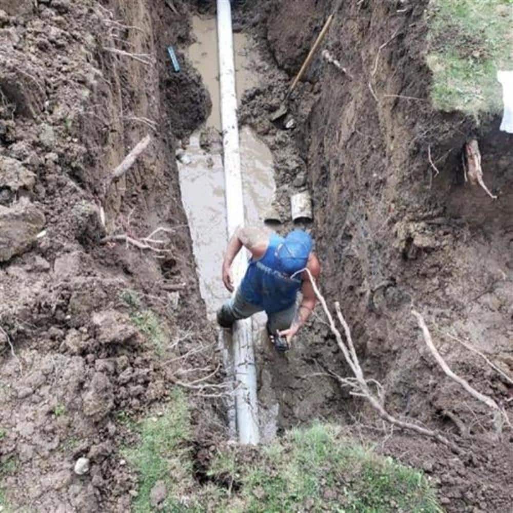 A Bison Plumbing technician working at the bottom of a deep trench beside a house, tree roots protruding from the clay trench walls around the new white PVC sewer lateral