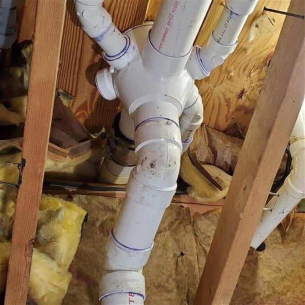 A newly installed white PVC drain assembly with wye fittings and cleanout running between floor joists and insulation in a Macomb County basement ceiling