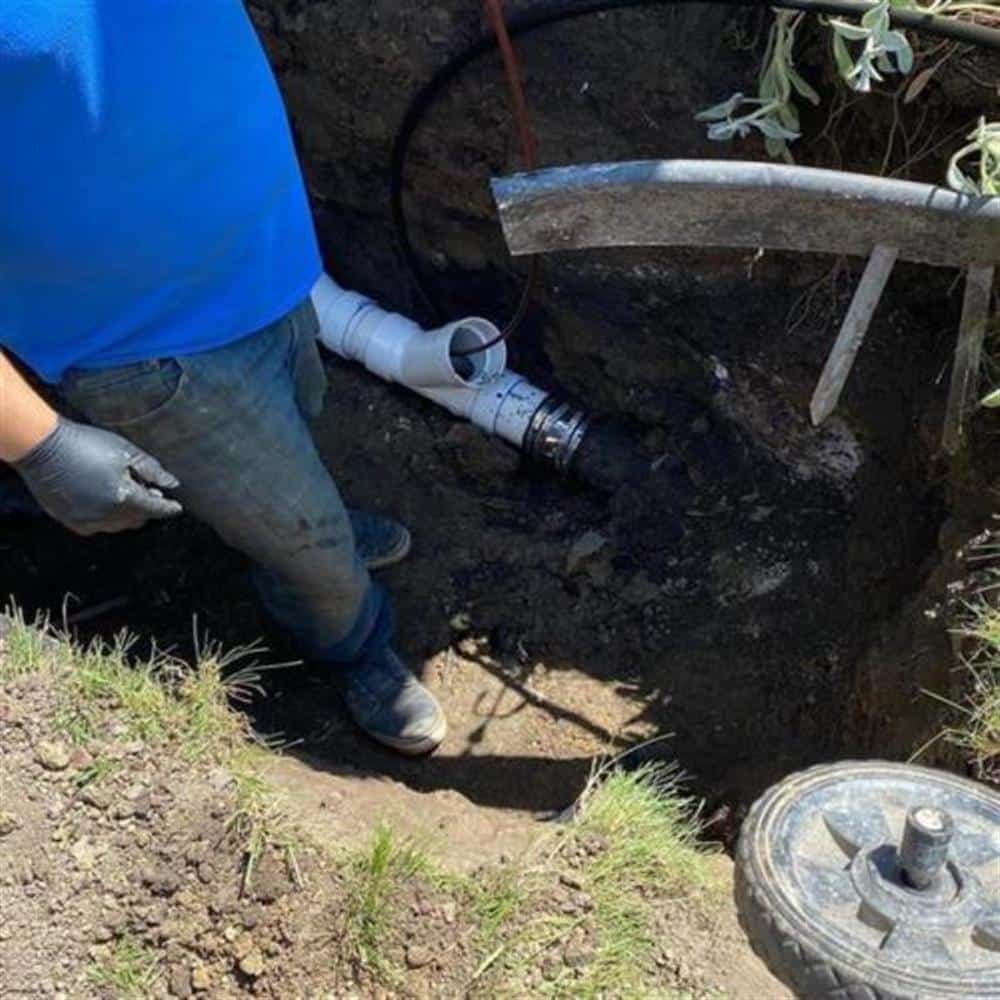 A Bison Plumbing technician kneeling at a small excavation pit in a lawn, joining new white PVC to the existing sewer line with a banded coupling