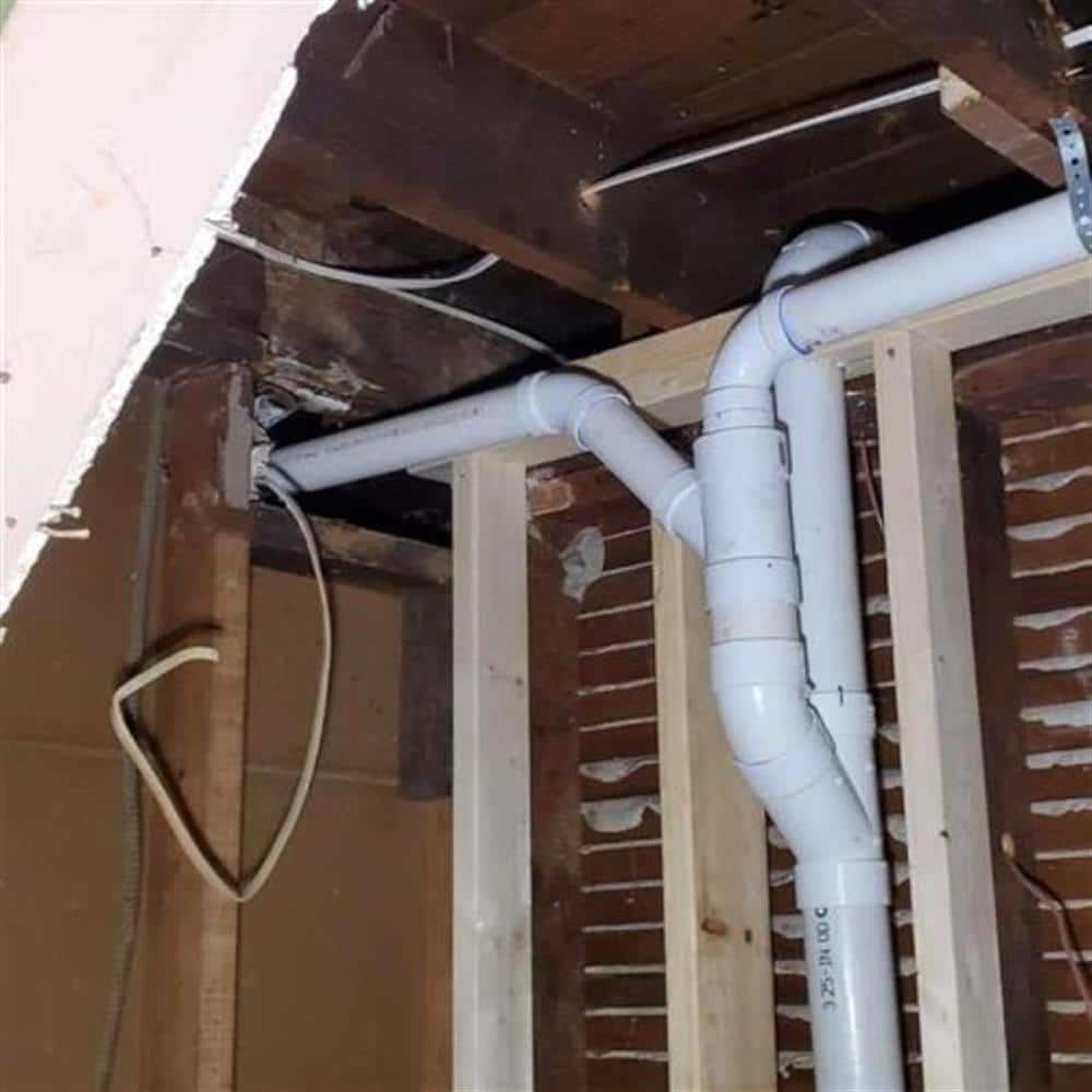 New white PVC branch drain lines running at a consistent slope between exposed floor joists and wall studs in a basement remodel