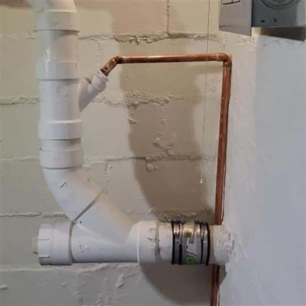 A white PVC drain line with a cleanout fitting and shielded coupling running down a painted block basement wall beside a copper water line