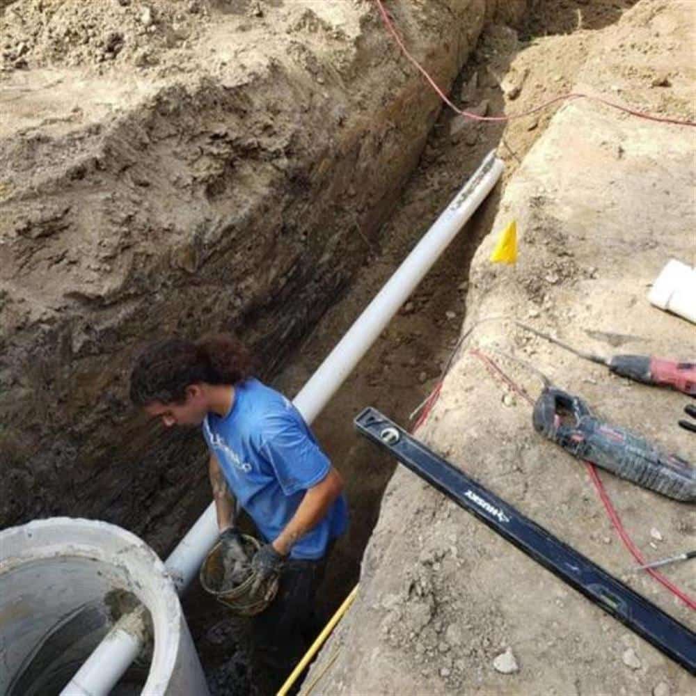 A Bison Plumbing technician kneeling in a compact access pit in a lawn, guiding a section of white PVC into the exposed sewer lateral with a level and tools laid out on the grass