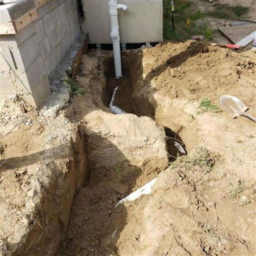 A small, neat access pit dug along a house foundation by Bison Plumbing, with the white PVC sewer lateral exposed at the bottom and a shovel resting on the lawn beside it