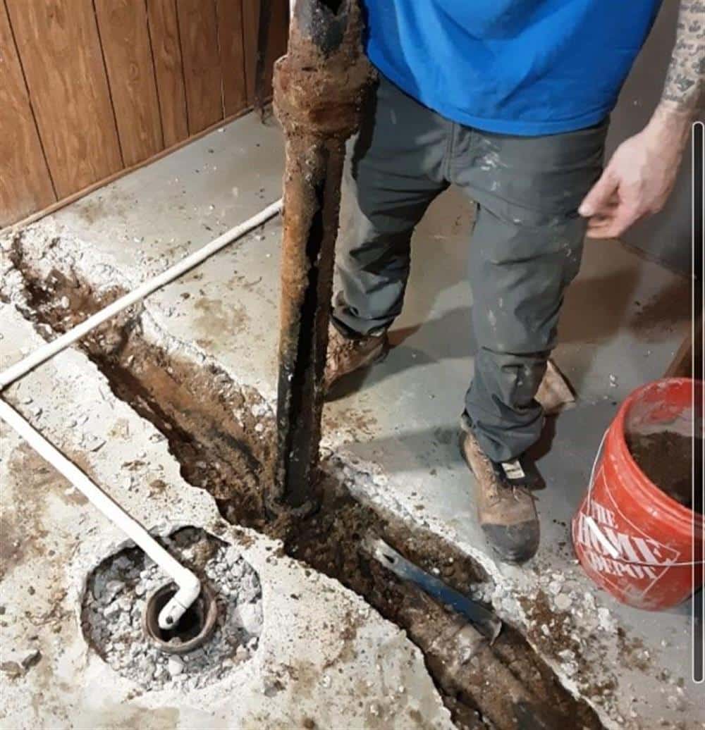 A corroded, broken cast-iron drain pipe lifted out of a trench cut through a basement floor, with a Bison Plumbing technician's boots and a red bucket beside it