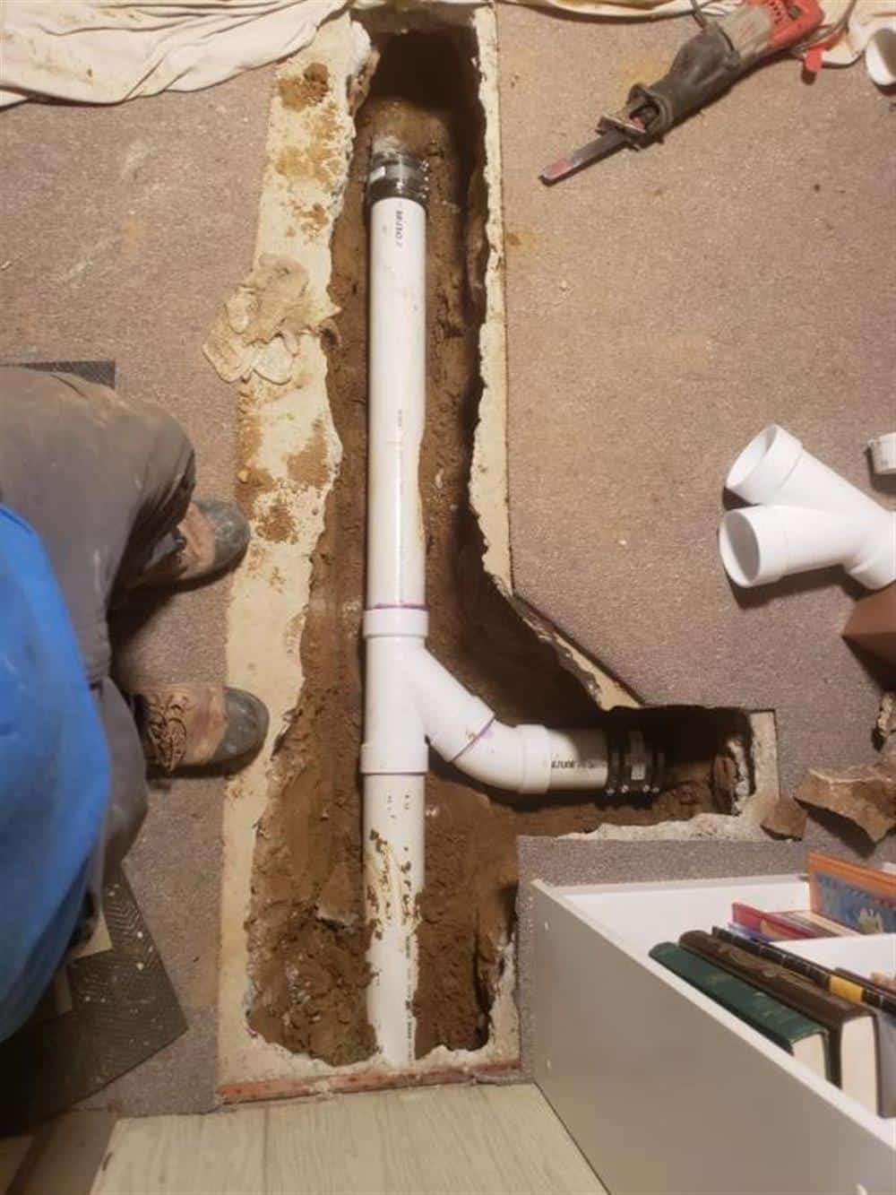 A Bison Plumbing technician kneeling at a trench in a basement floor, setting a new white PVC drain fitting with fresh cement around it, spare fittings on the floor nearby