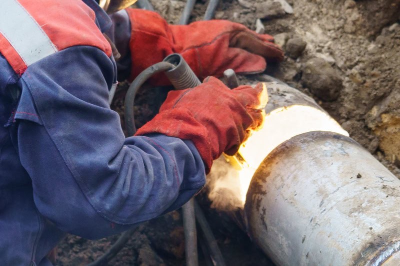 A worker wearing protective gloves and clothing welds a metal pipe outdoors, with bright sparks at the welding point. Cables and soil surround the work area—just another day for expert plumbers in Warren MI handling trenchless sewer repair.