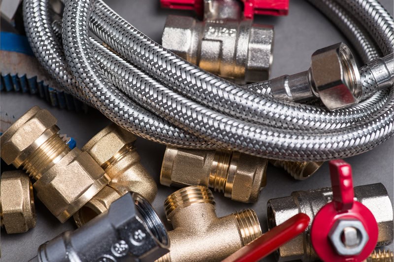 Assorted metal plumbing fittings, braided hoses, and red-handled valves are shown close up, arranged together on a gray surface—perfect supplies for plumbers in Warren MI or anyone looking for reliable solutions like trenchless sewer repair.