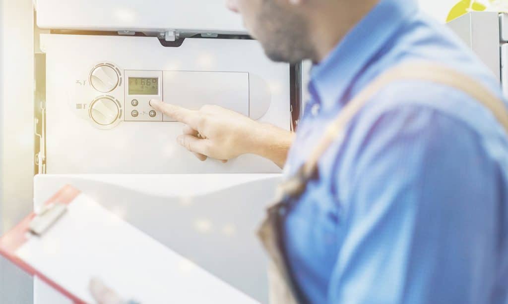A bison plumbing technician in a blue shirt and overalls uses a digital control panel on a white heating system, holding a red clipboard—your trusted "plumber near me" for efficient service.