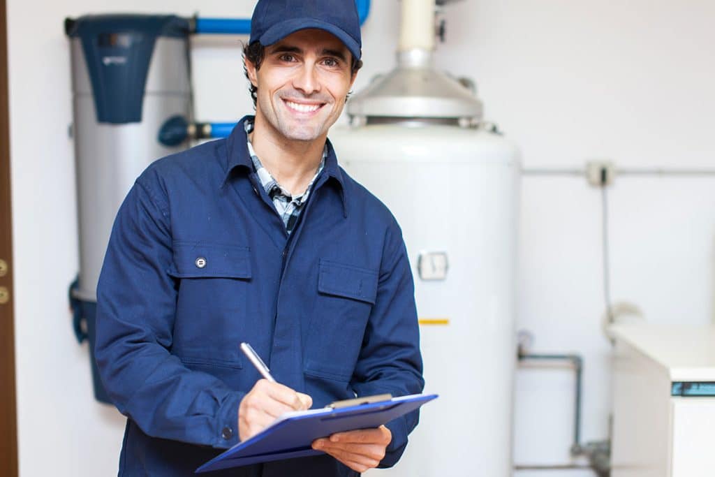 A smiling maintenance worker in a blue uniform and cap holds a clipboard and pen, standing in front of water heaters and pipes—ideal for anyone searching "plumber near me" or needing help from Bison Plumbing Warren Michigan.
