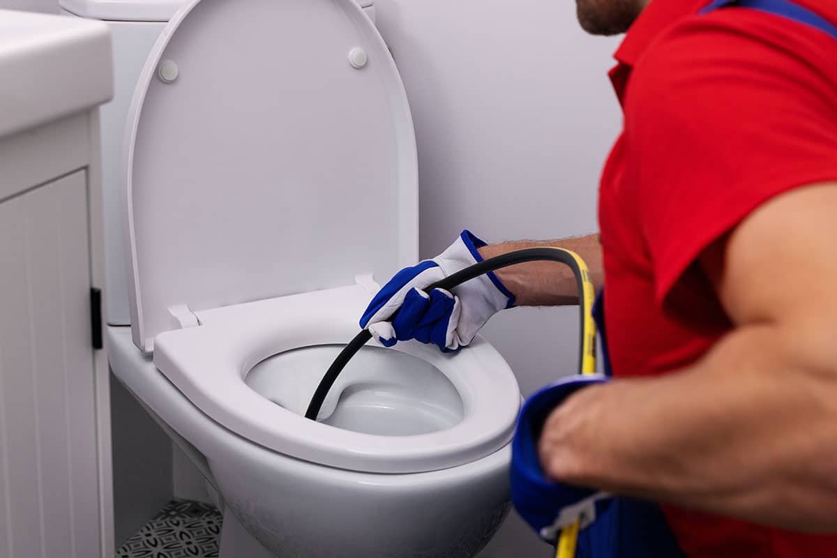 A person wearing gloves and a red shirt uses a plumber’s snake tool to unclog a white toilet in a bathroom, showcasing the expertise of plumbers in Warren MI from bison plumbing.