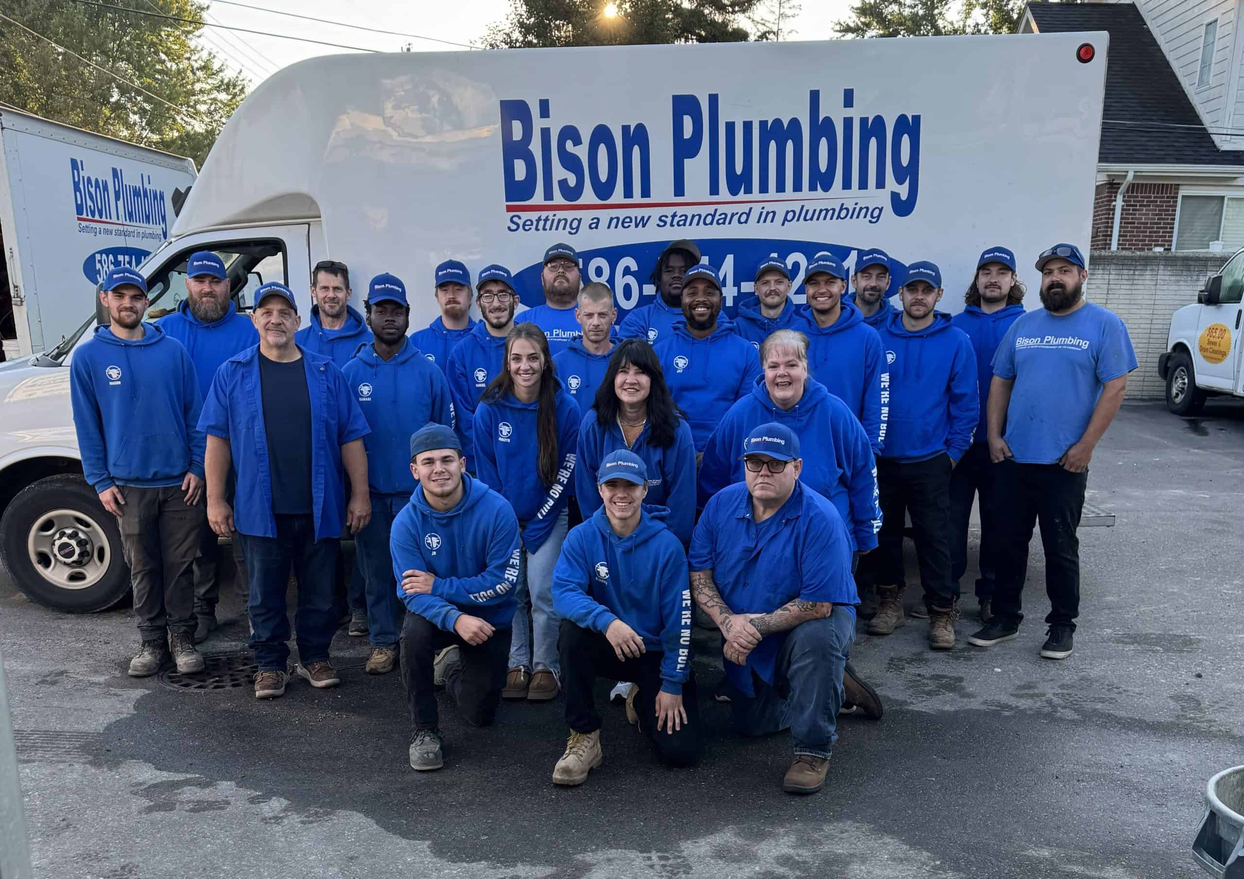A group of 20 people wearing blue Bison Plumbing shirts and hats pose and smile together in front of a Bison Plumbing van with the company logo on it, outside on a paved lot.